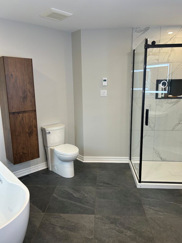Salle de bains moderne : toilettes, douche avec cadre noir, sol carrelé foncé, armoire haute en bois, murs gris clair.