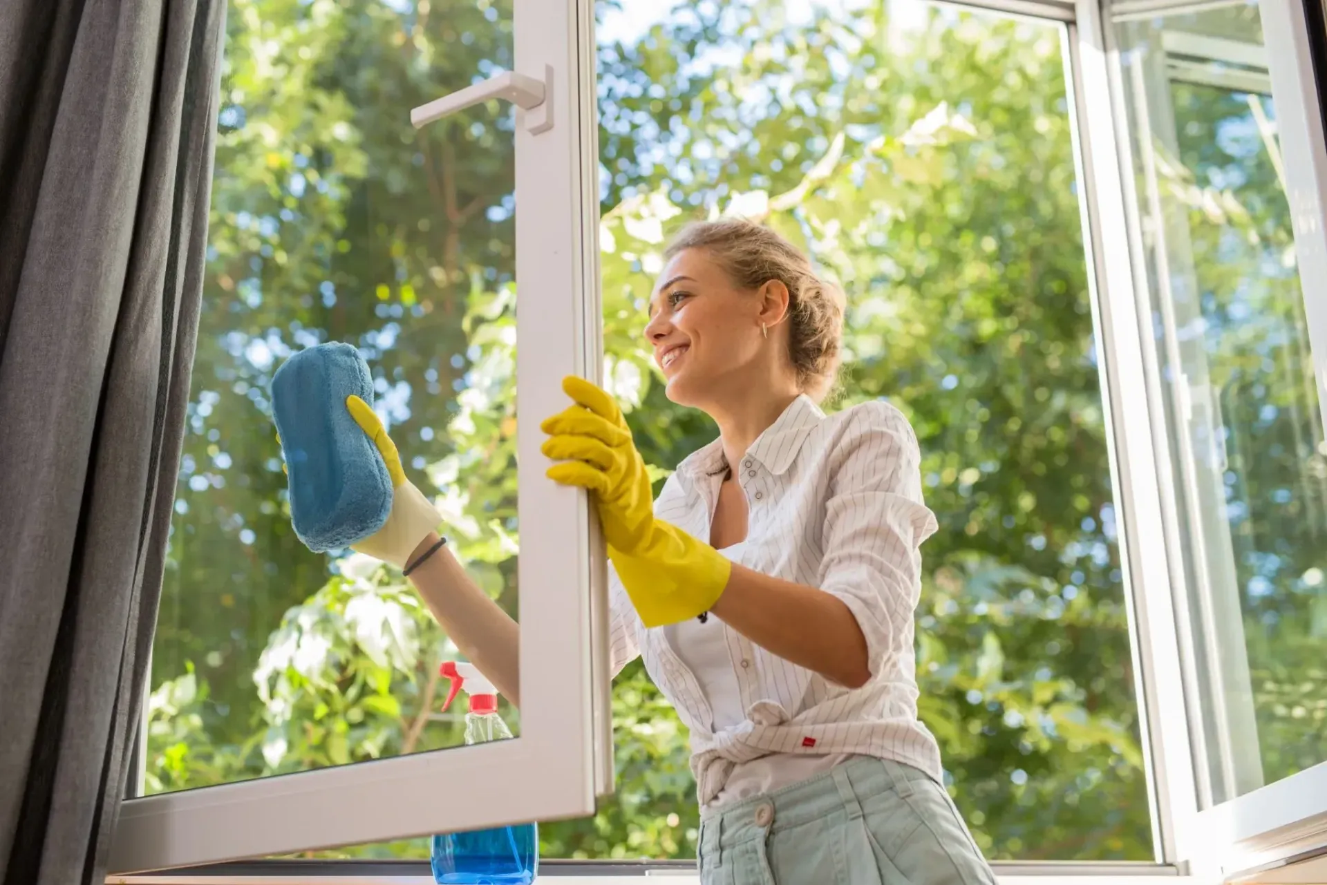 Woman in yellow gloves cleaning a window with a blue cloth in a bright room