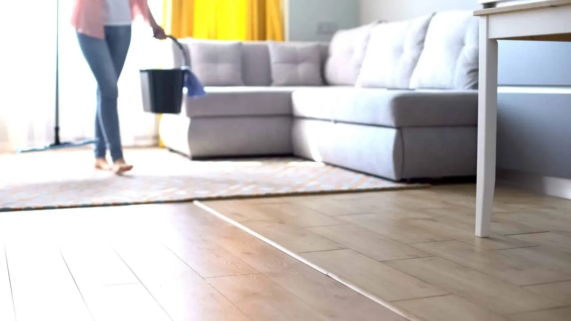 A bright living room with a gray sectional sofa, wooden floor, and a person walking in from the left carrying a black bucket.