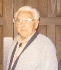 An older person wearing glasses and a light-colored cardigan over a shirt with a bolo tie, standing before wooden doors.