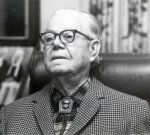 A person with glasses, wearing a checkered jacket and a bolo tie, seated against a neutral background.