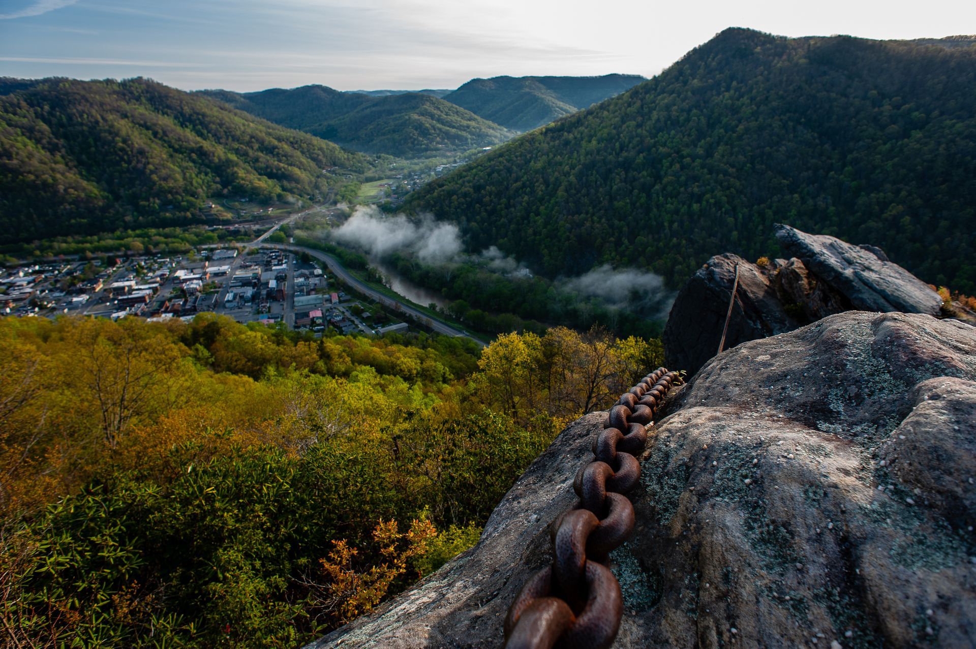 A heavy iron chain rests on a rock overlook, featuring a panoramic view of a valley town nestled between green mountains.