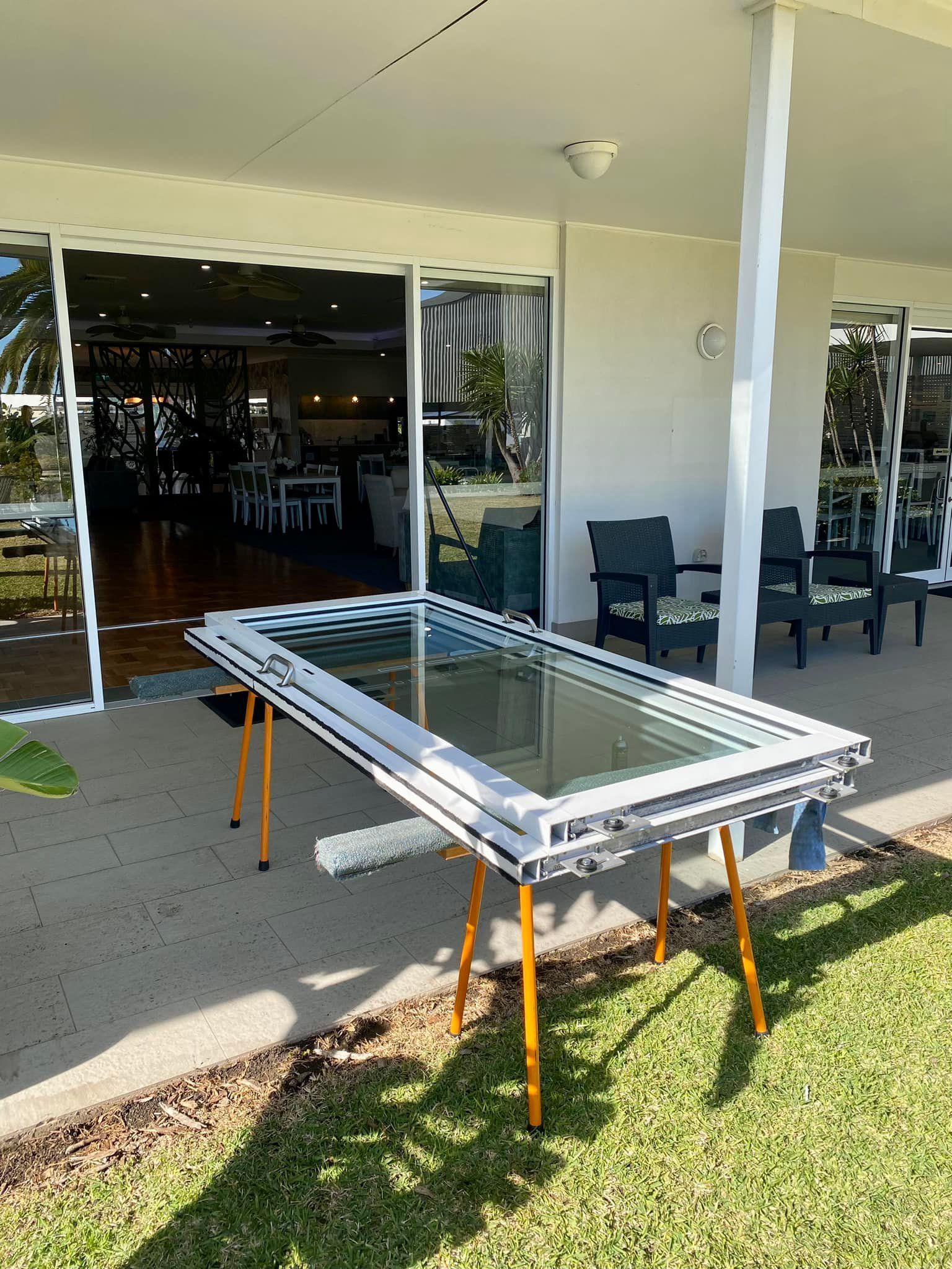 A Window Frame With Glass Sits on Sawhorses Outside a Building With Sliding Glass Doors — Fix-A-Door in Lake Macquarie, NSW