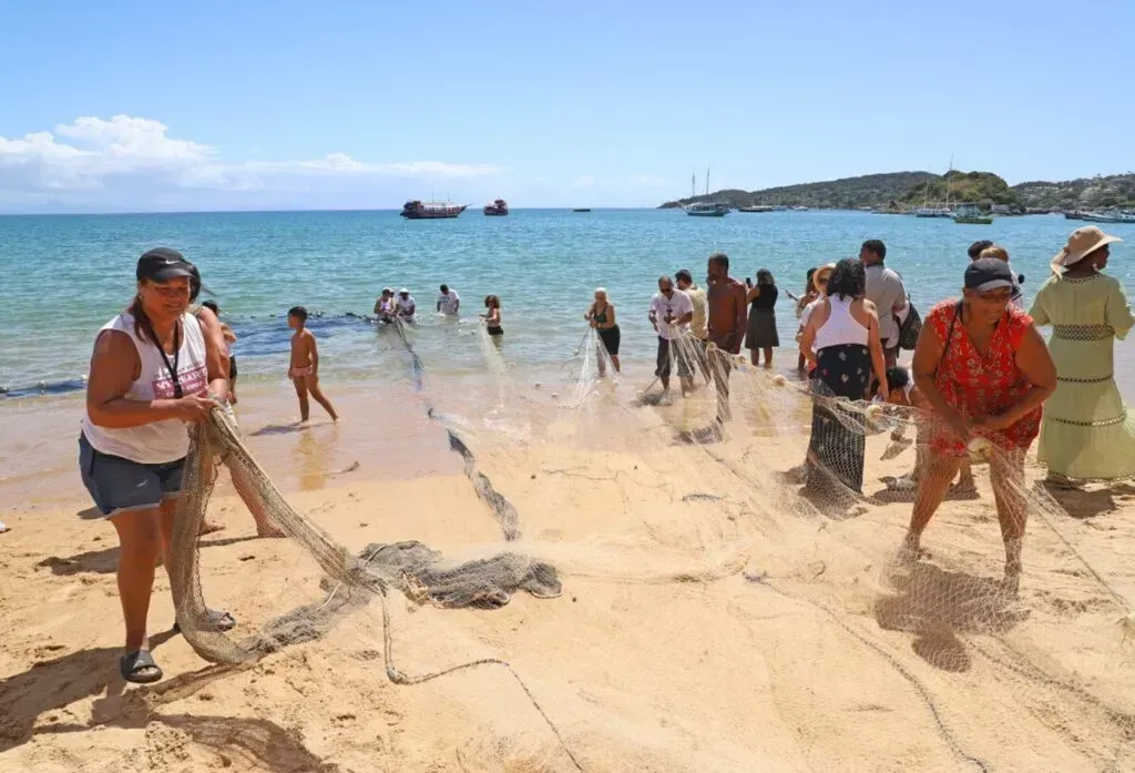 Pessoas puxando uma rede de pesca do oceano para uma praia de areia sob um céu azul.