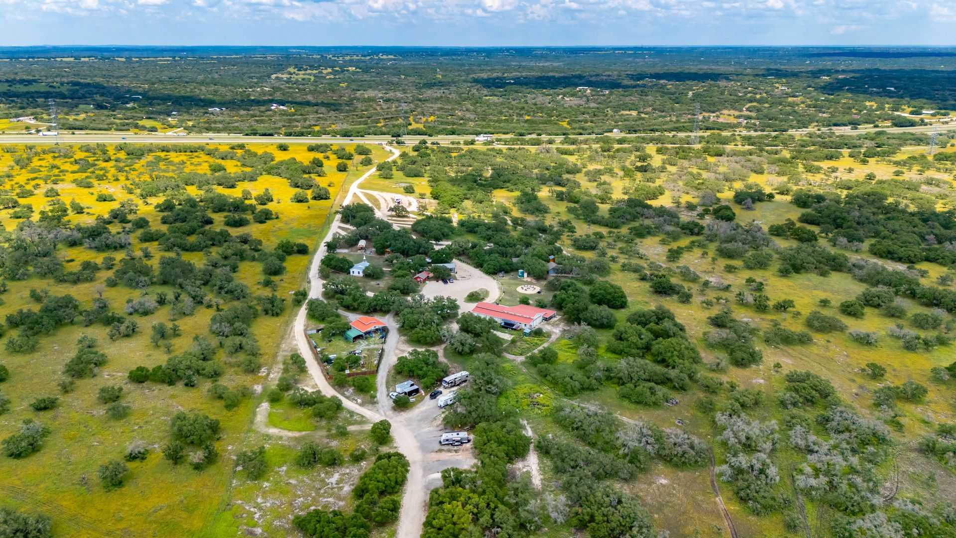 Aerial view of a green rural landscape with scattered trees, dirt roads, and a small cluster of buildings.