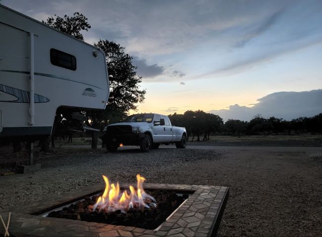 Campfire glowing at dusk beside a white RV and pickup truck in a campsite.