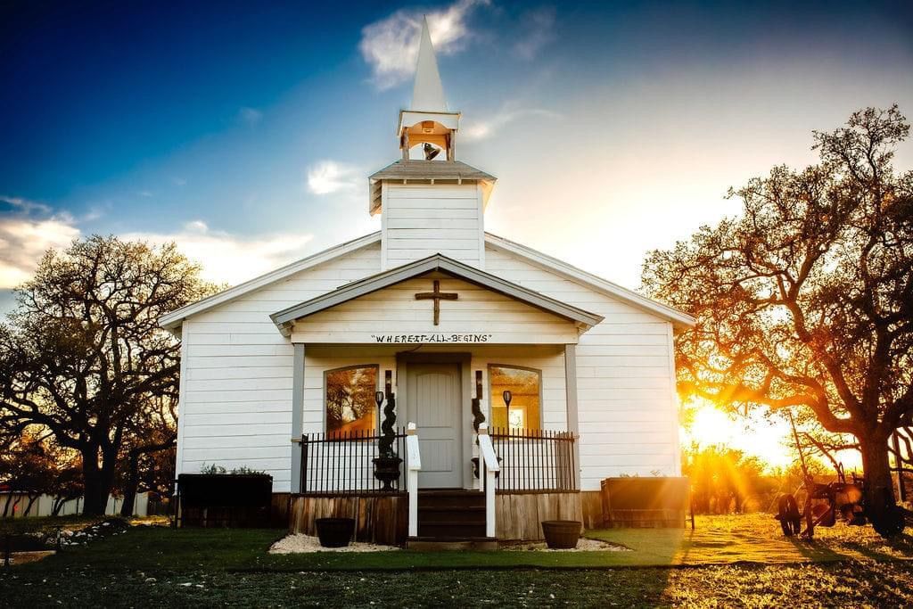 Small white church at sunset with a steeple, framed by bare trees and warm golden light
