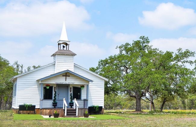 Small white rural church with steeple and porch, surrounded by grass and trees under a blue sky.