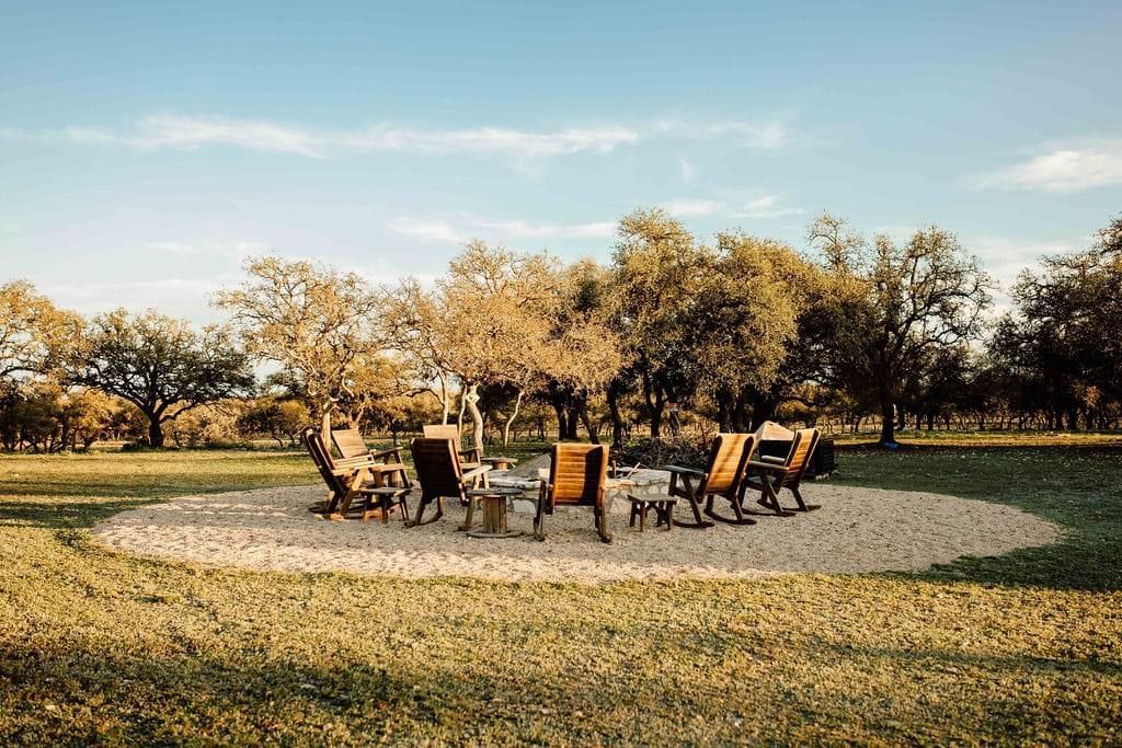 Outdoor fire pit with chairs arranged on a grassy field under trees at sunset