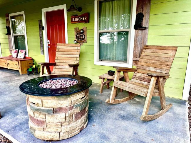 Front porch with two wooden rocking chairs, a round fire pit, and a red door on a lime-green house.
