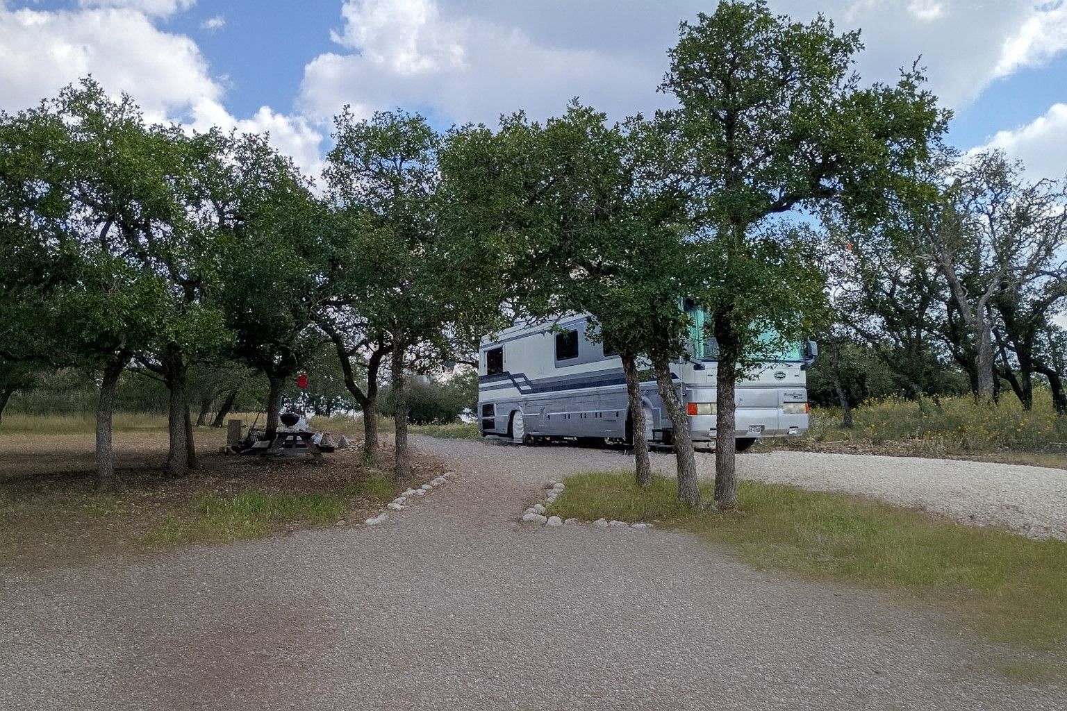 RV parked under trees on a gravel lot with a cloudy sky overhead
