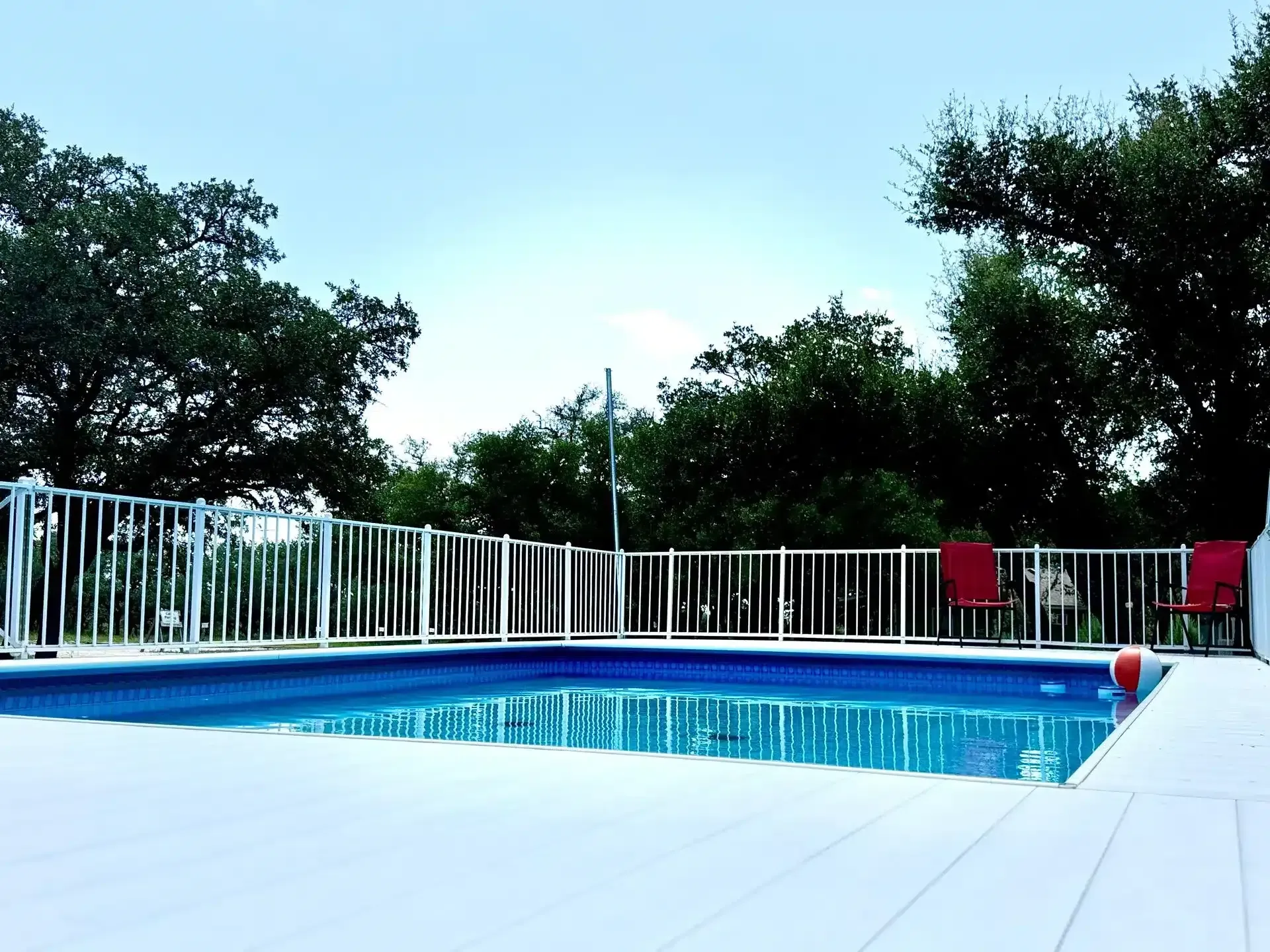 Backyard pool with white deck, white fence, and two red lounge chairs under trees