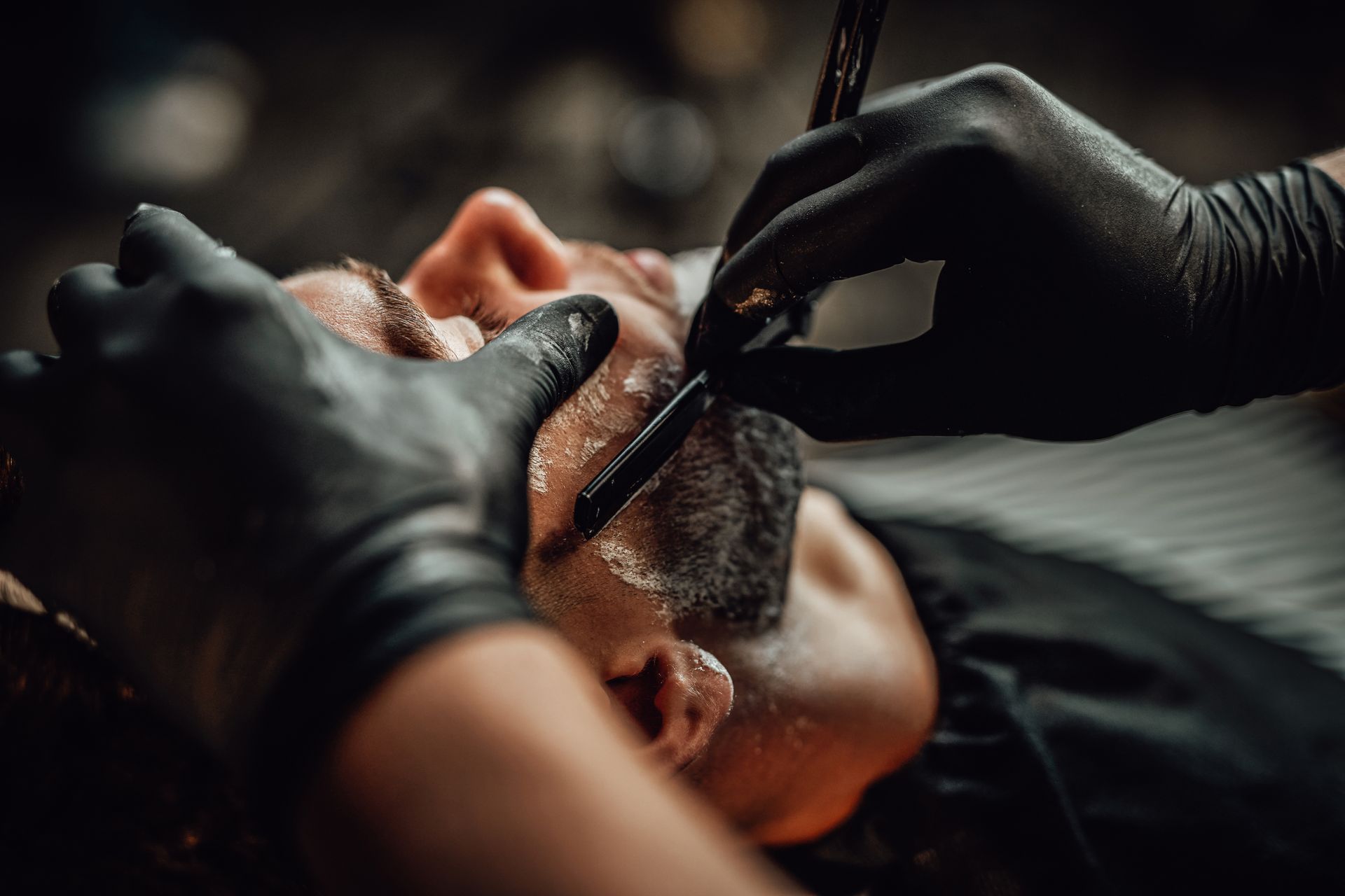 A man is getting his beard shaved by a barber wearing black gloves.