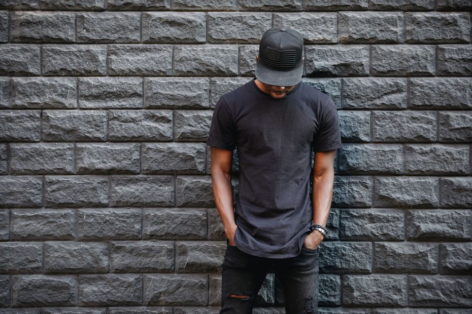 A man wearing a black hat and a black t-shirt is standing in front of a brick wall.