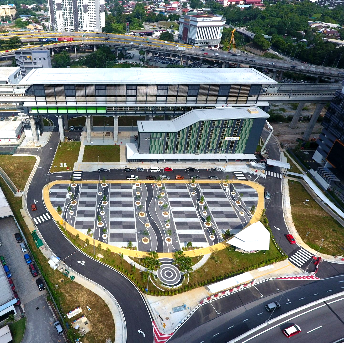 Elevated train station with parking lot, curved road, and surrounding buildings.