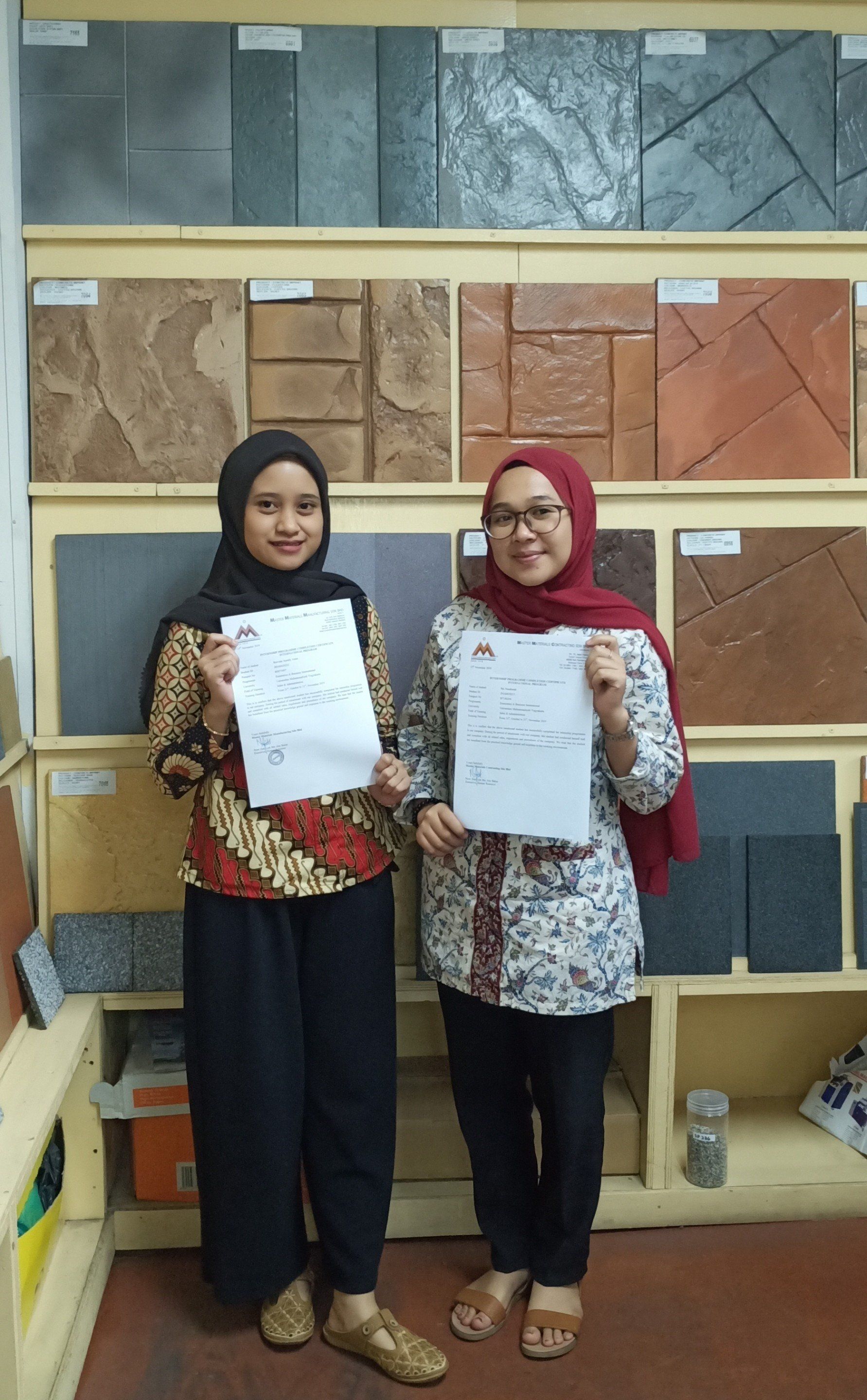 Two women smiling, holding papers, standing in front of a tile display.