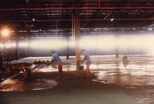 Two workers smoothing concrete in a large, empty warehouse.