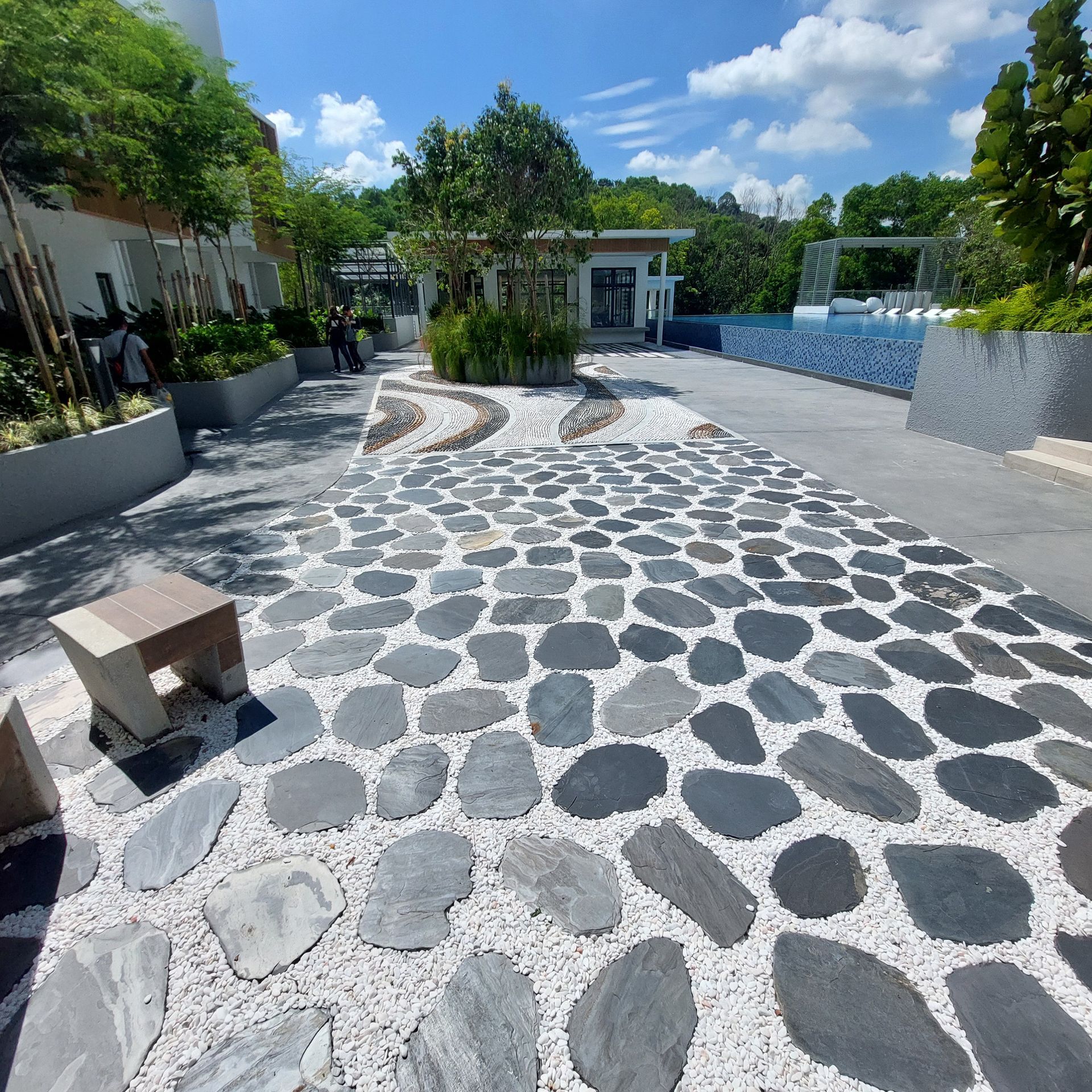 Path paved with gray stones and white pebbles, leading to building with a pool, trees and blue sky.