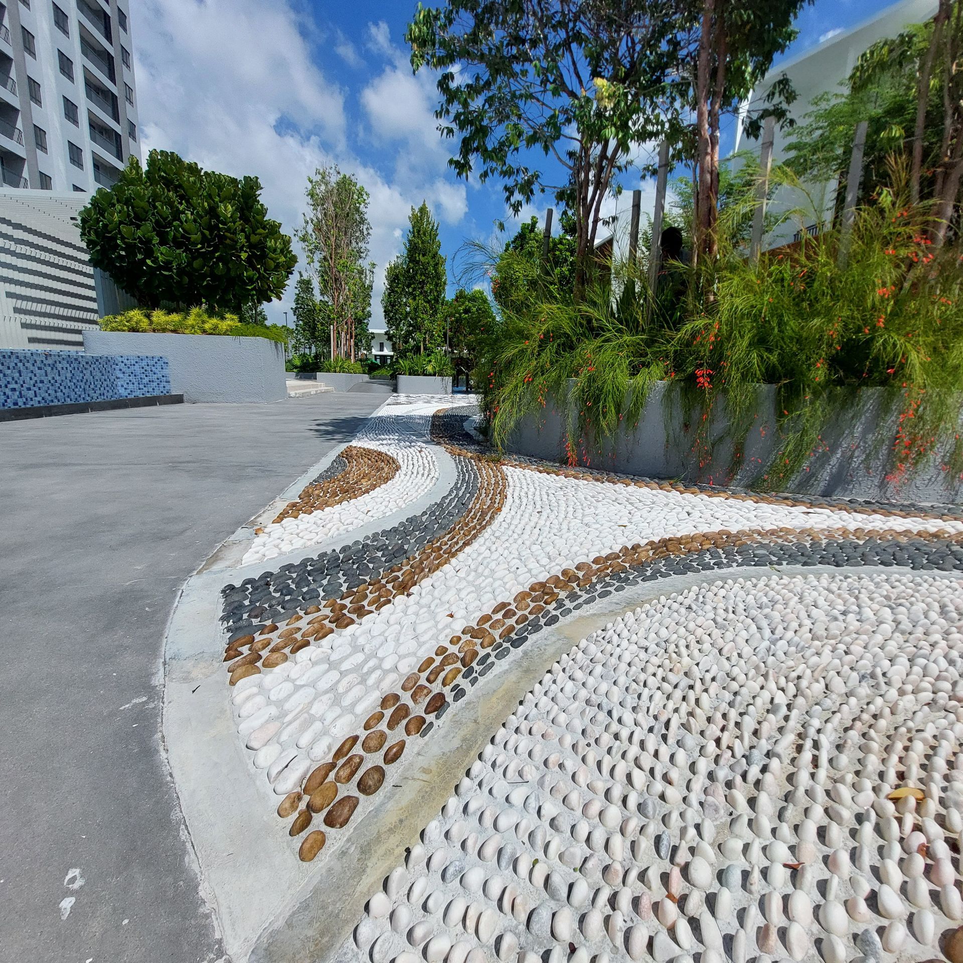 Pebbled path with flowing design, bordered by concrete and landscaping, under a partly cloudy sky.