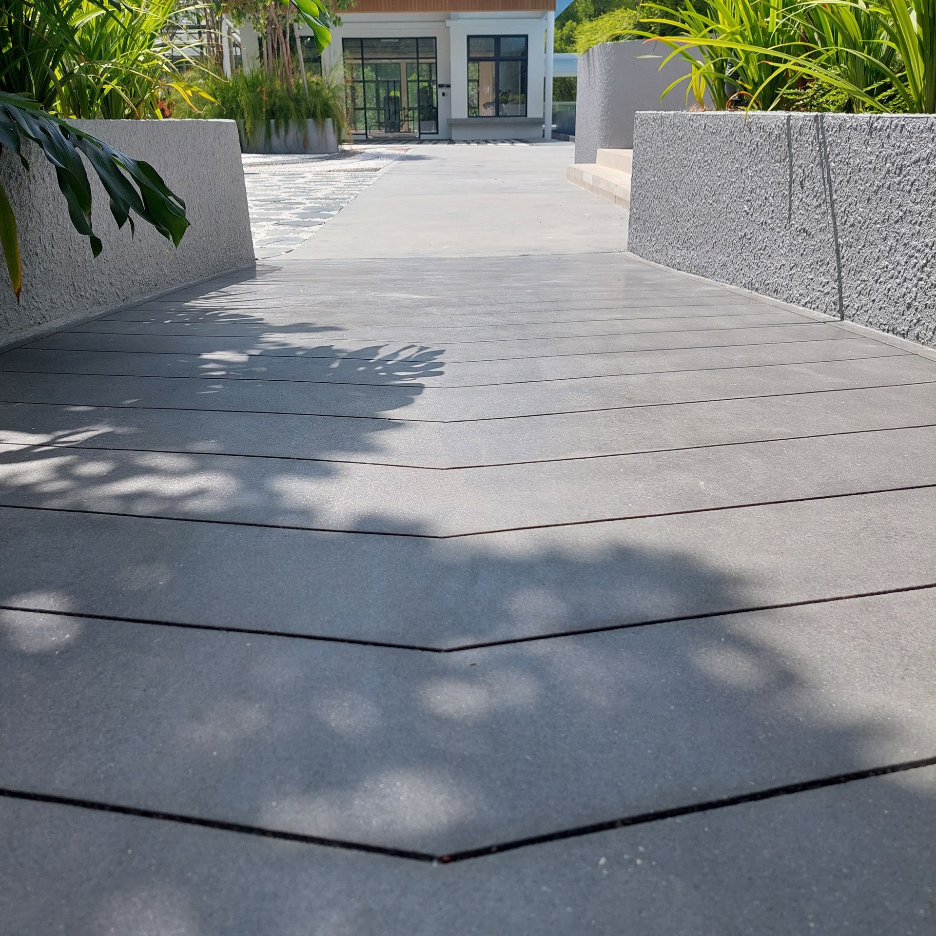 A gray walkway with textured walls leads to a building, surrounded by greenery.