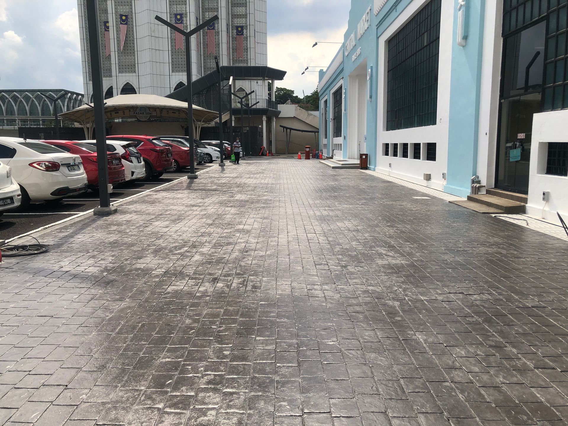 Brick-paved walkway with cars parked on the left and a light blue building on the right, cloudy sky.