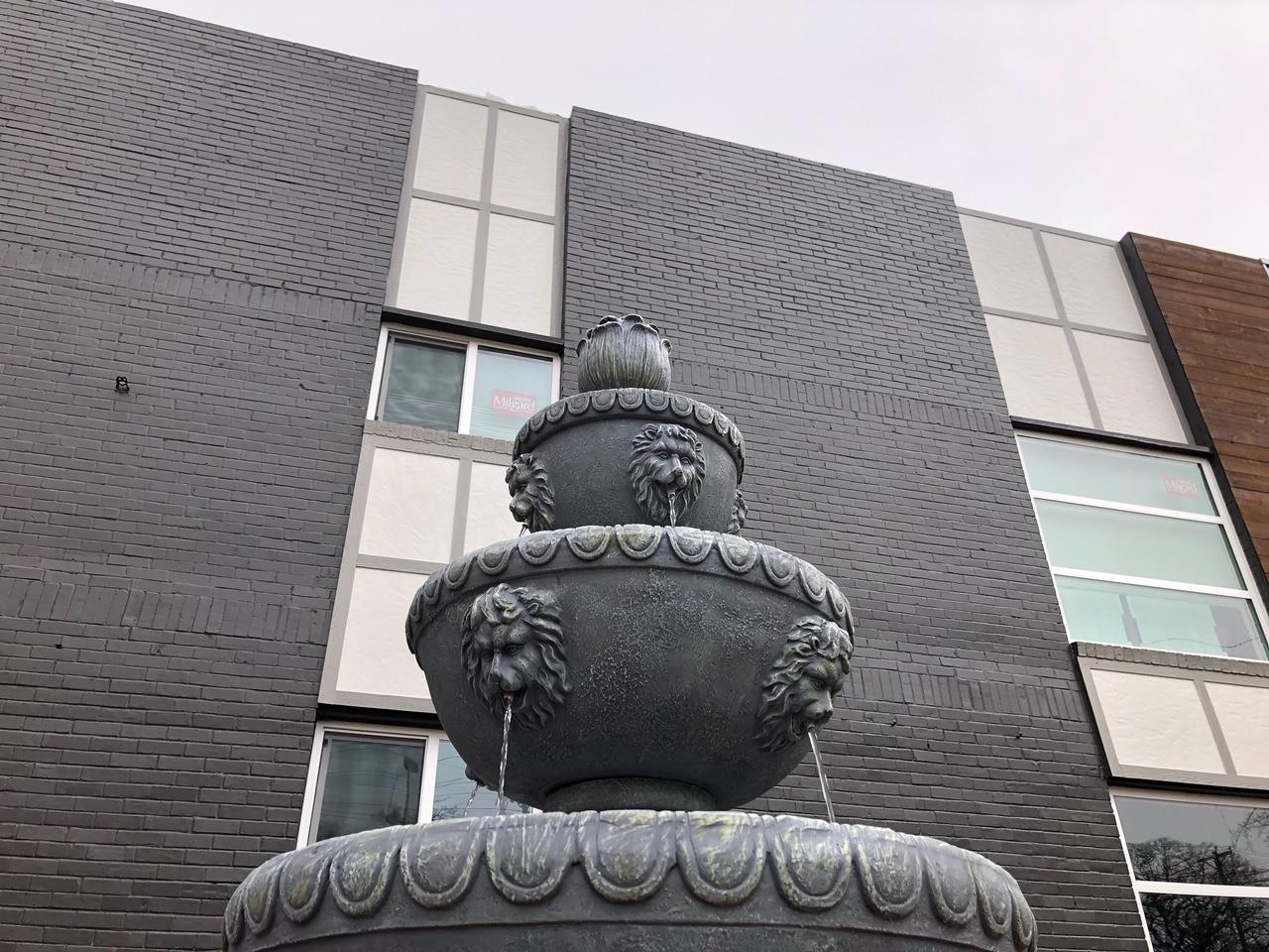 Stone fountain with lion head spouts in front of a modern building with windows and dark gray textured walls.