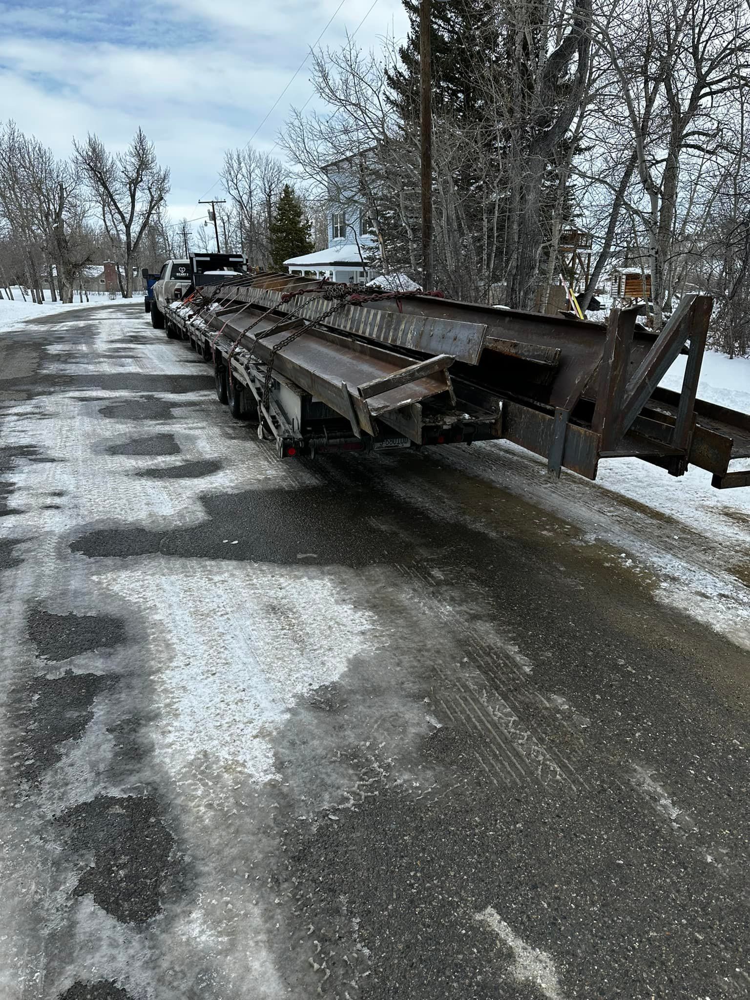 A long, rusty metal trailer sits on an icy asphalt road in a snowy outdoor setting.