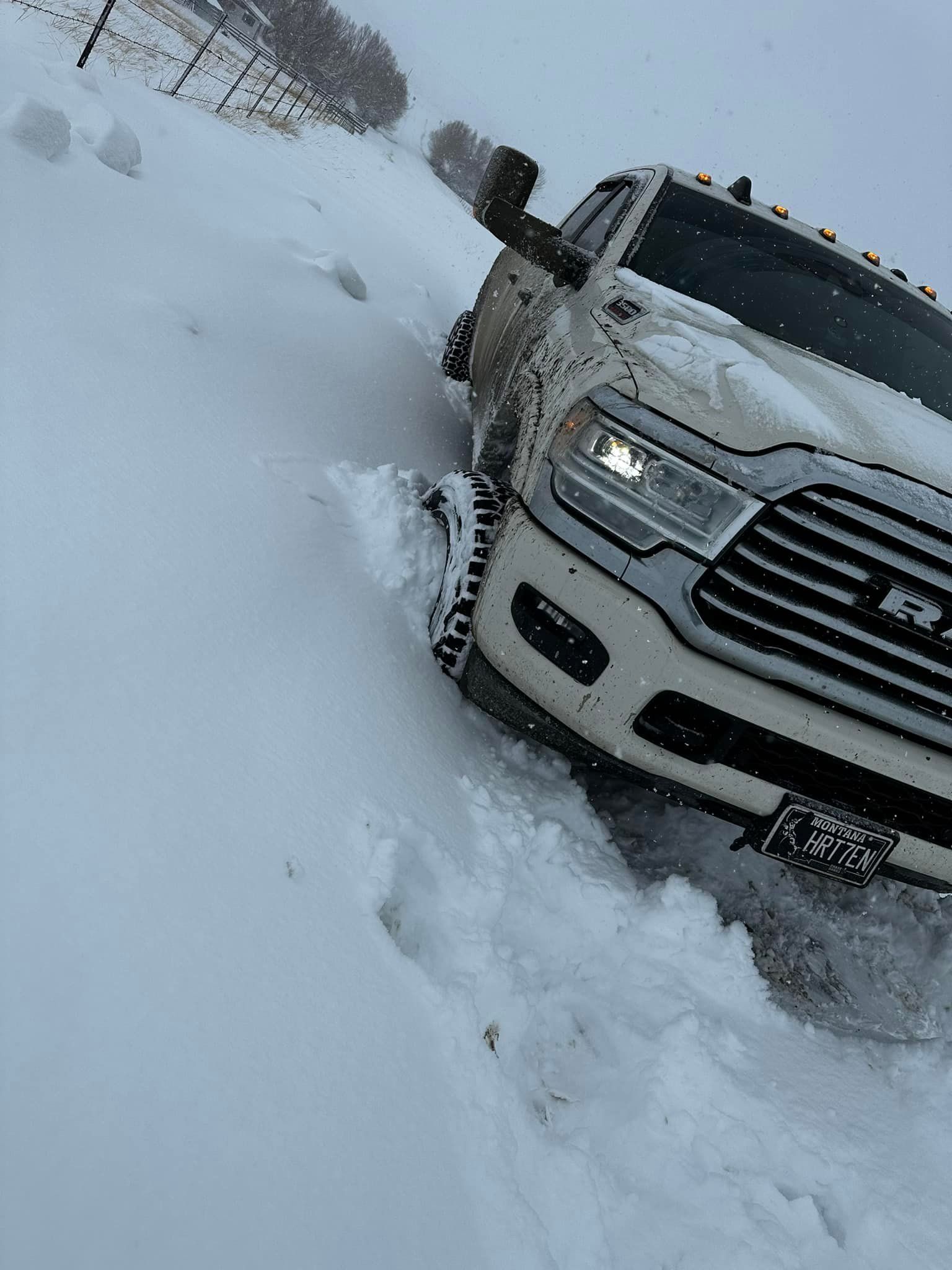 White pickup truck stuck in deep snow, front wheels buried. Snowy road and overcast sky.