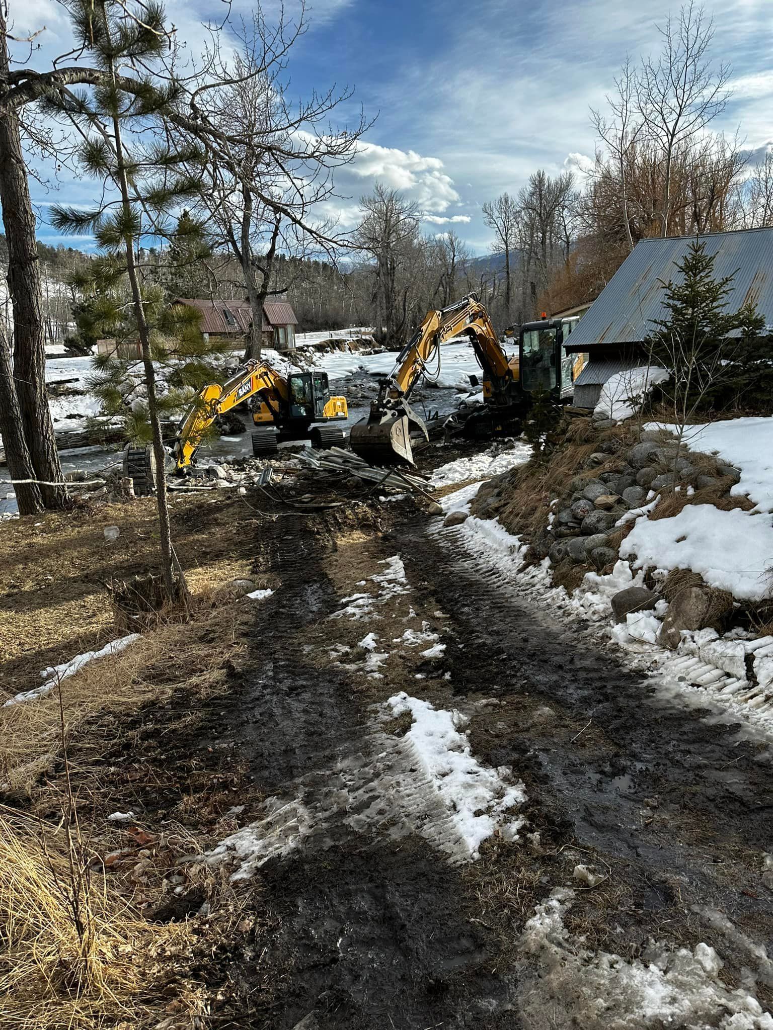 Two excavators digging near a building with snow on the ground; sunny day.