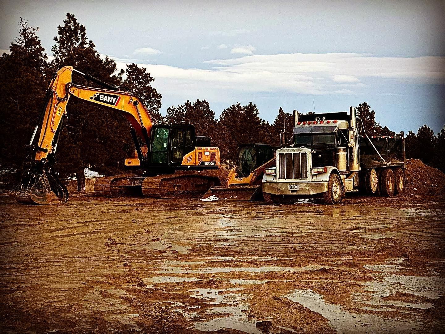 Excavator and truck on muddy ground in a construction site, trees in the background, overcast sky.