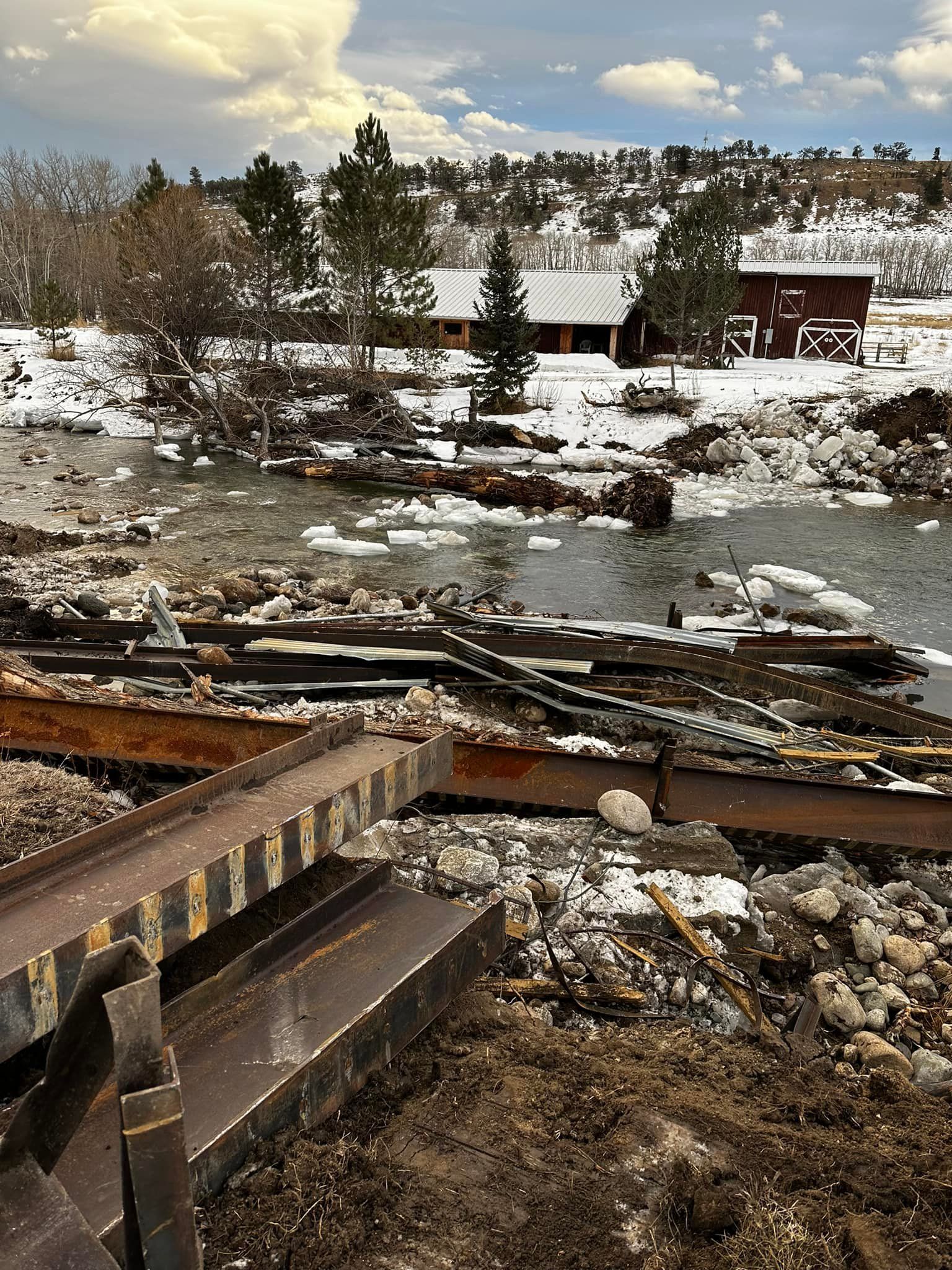 Snowy landscape with debris-filled water, houses in background, overcast sky.