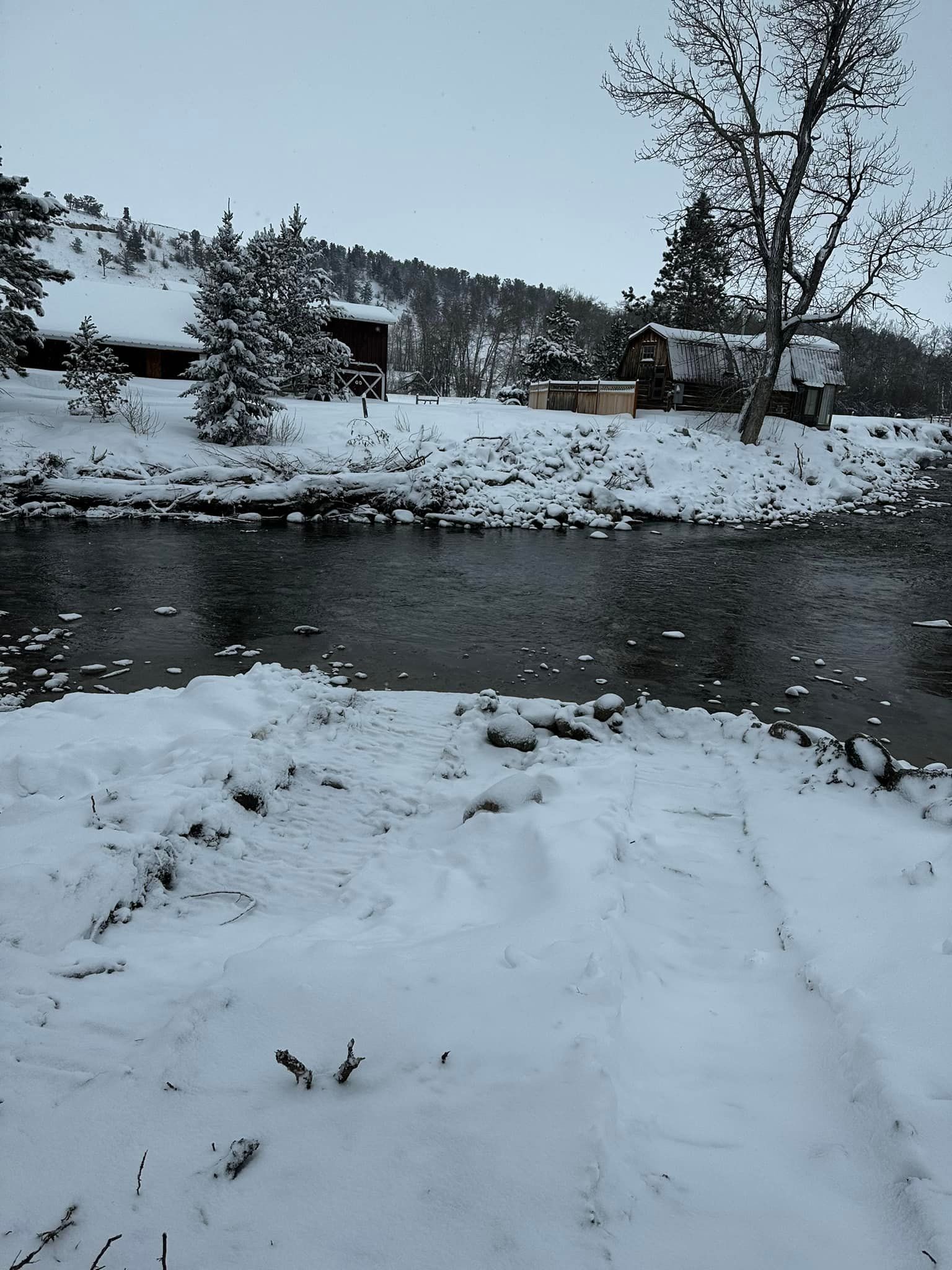 Snowy landscape with river, buildings, and trees under overcast sky.