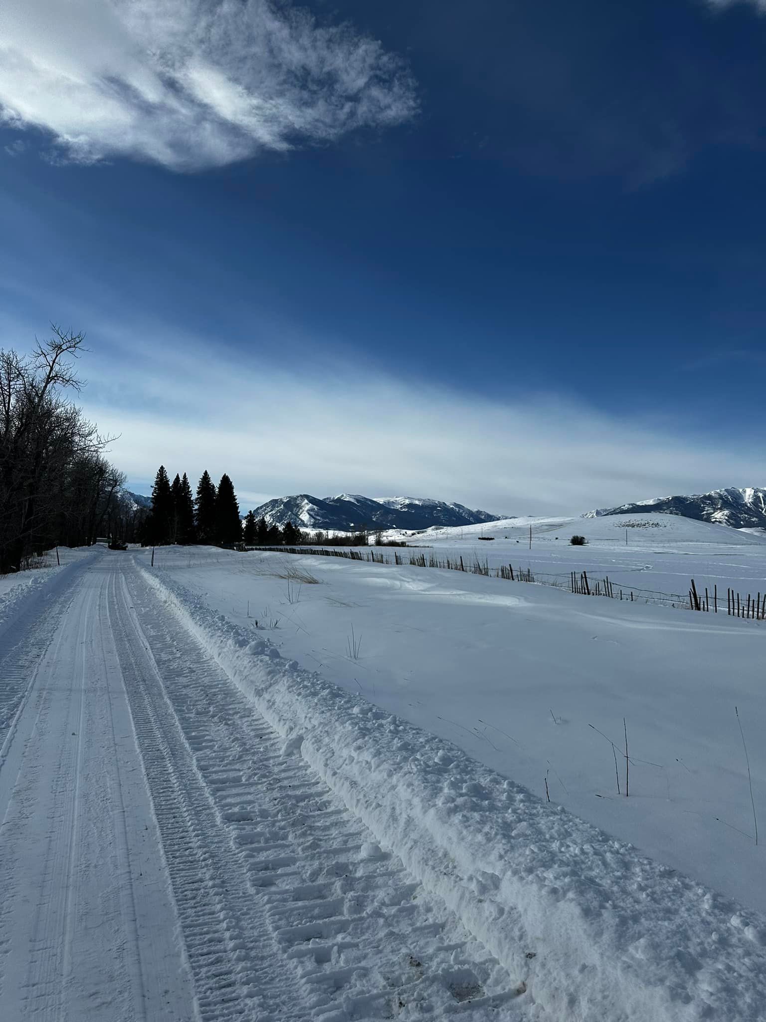 Snow-covered road leads towards distant mountains under a blue sky with some clouds.