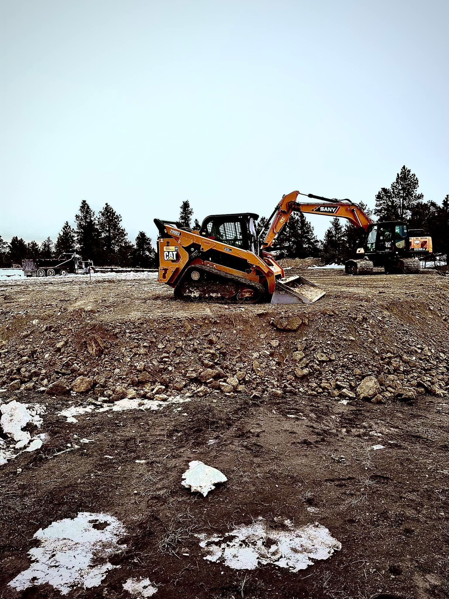 Two construction vehicles, an orange skid steer and excavator, on a gravel lot with snow patches.