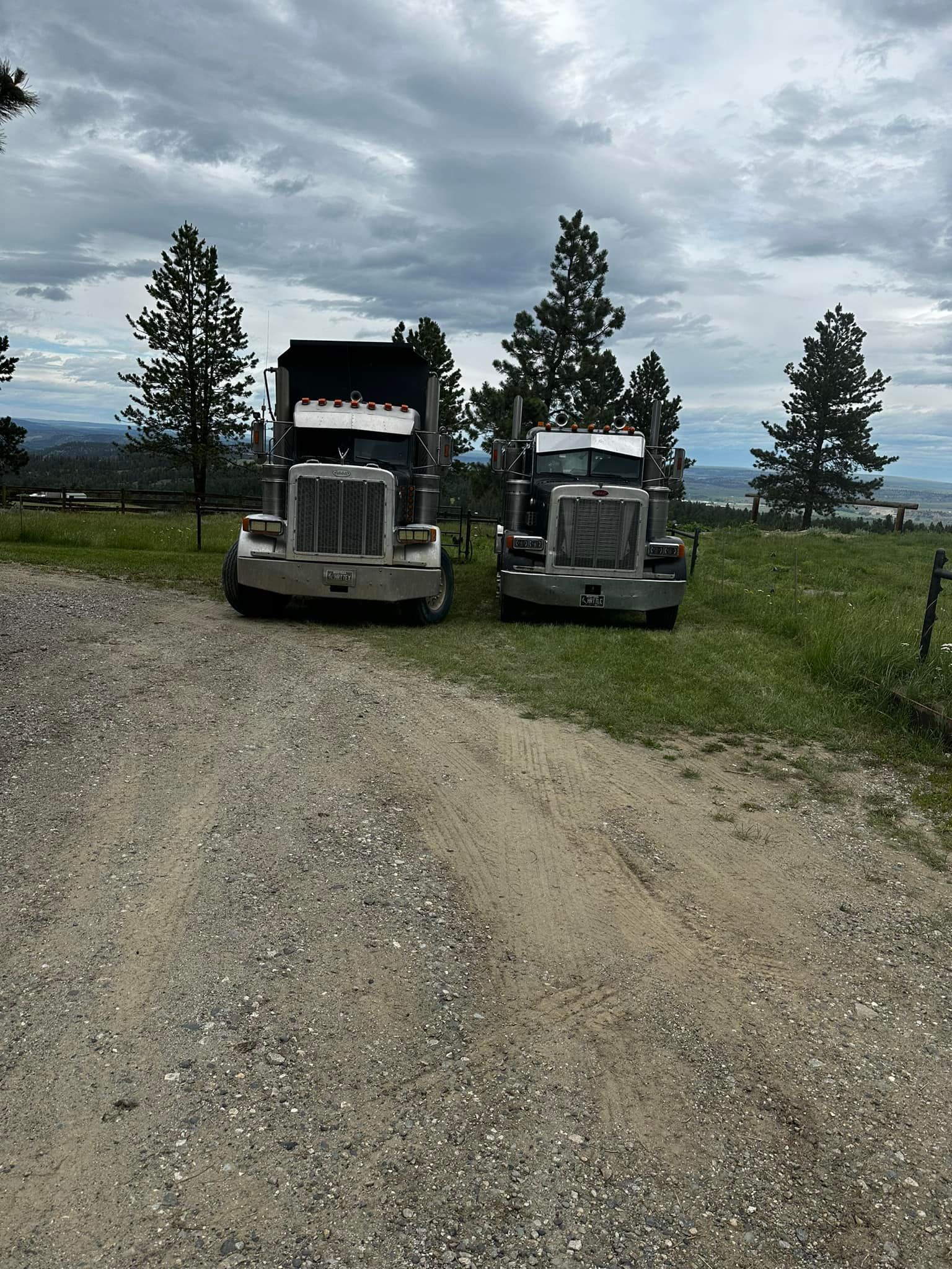Two semi-trucks parked on a gravel road, facing the camera. Cloudy sky, trees, and landscape in the background.