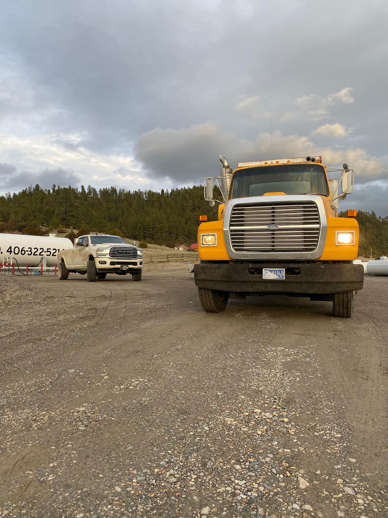 Yellow truck and white pickup on a gravel road, overcast sky, trees in the background.