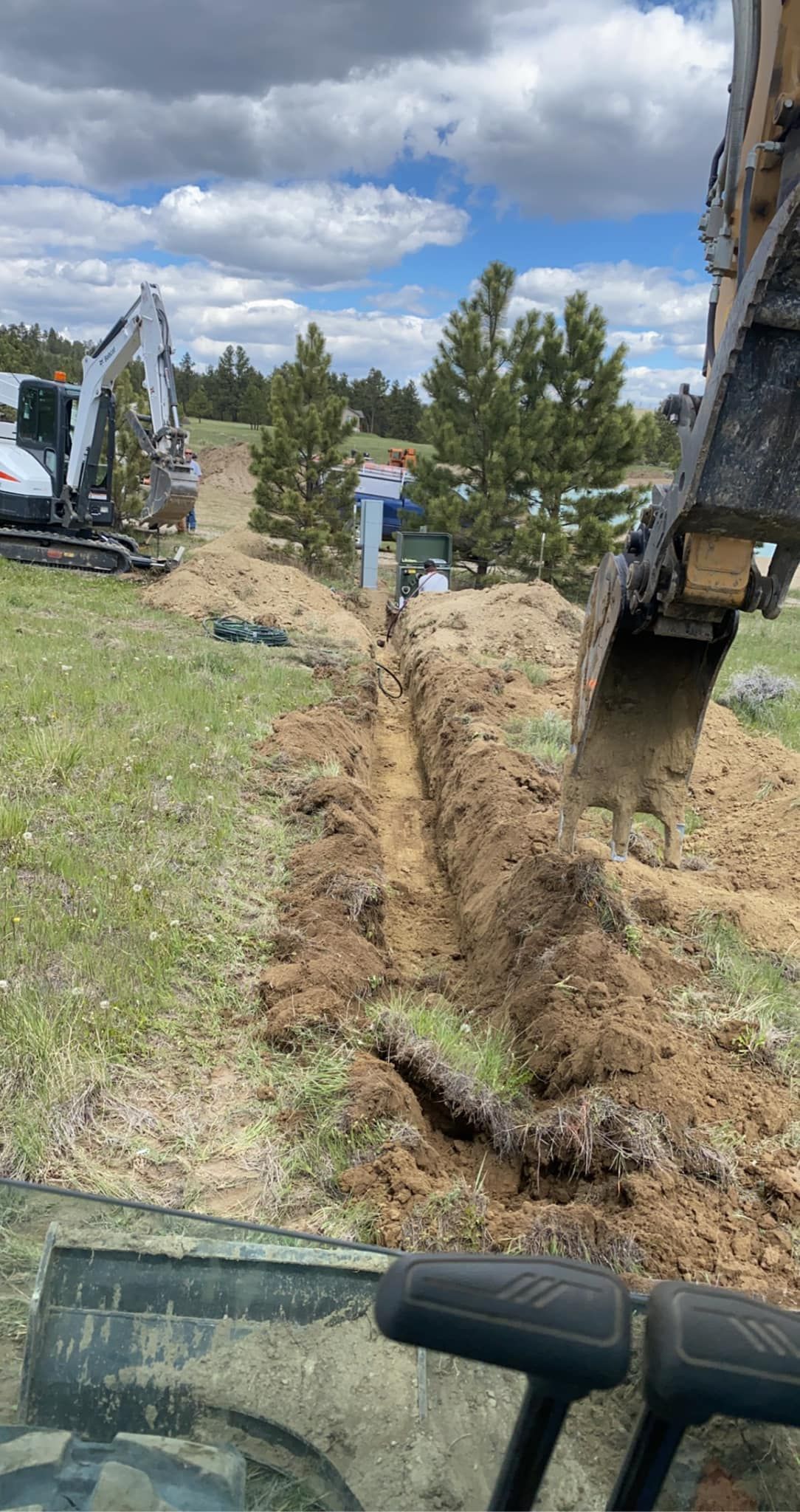 An excavator digs a trench in a grassy field, another excavator is in the background.