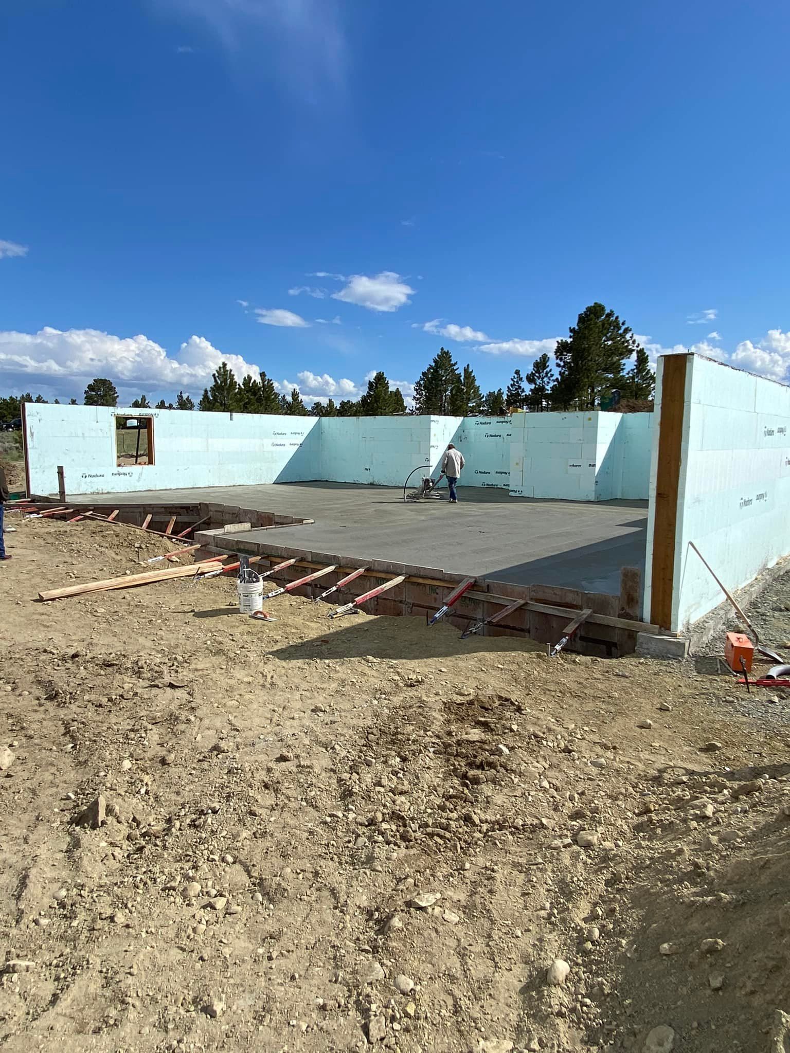 Construction site with blue foam walls, concrete floor, person working. Brown dirt ground, blue sky, trees.