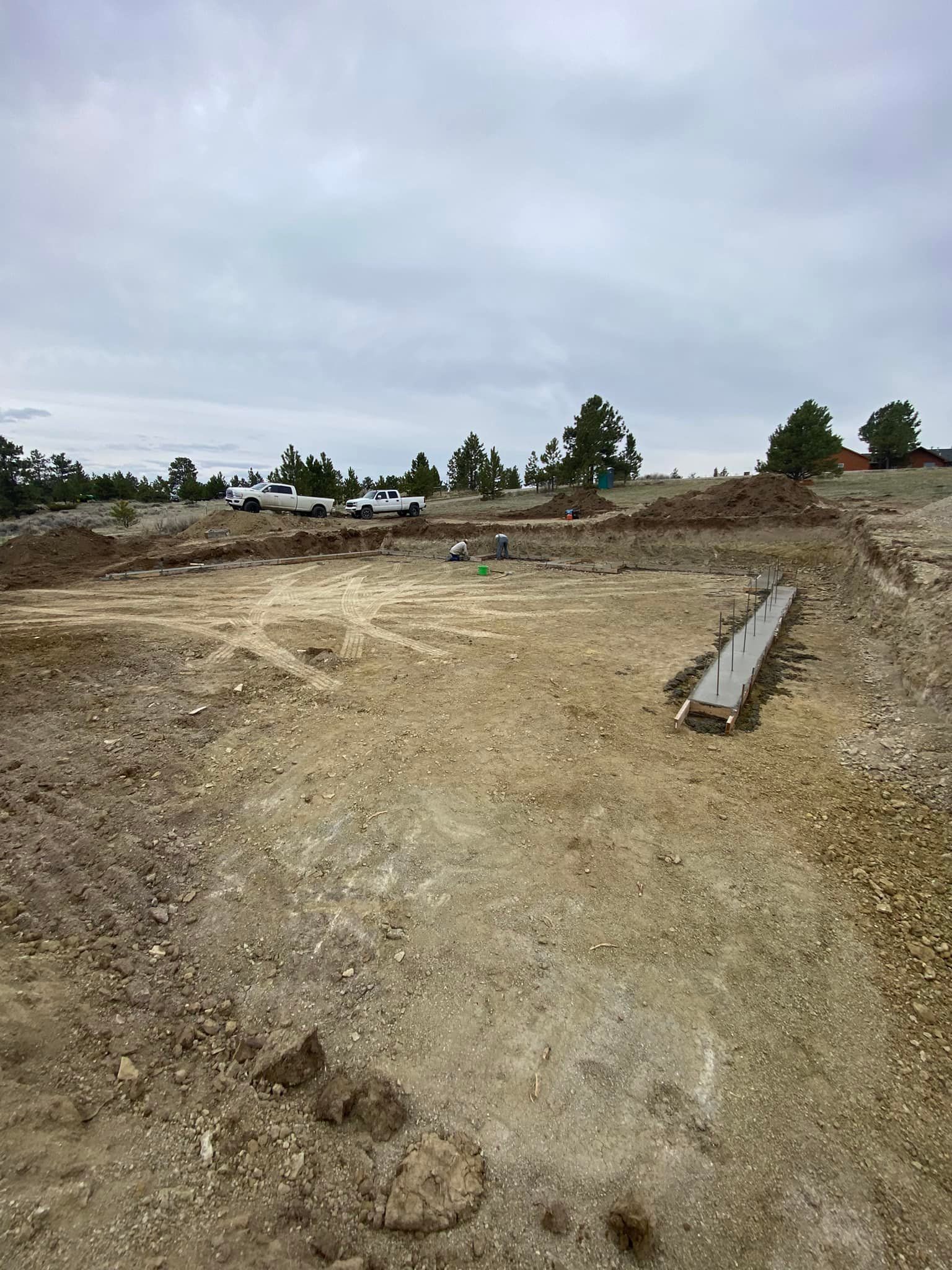 Construction site with dirt, gravel, and a wooden form. Vehicles and trees are visible under a cloudy sky.