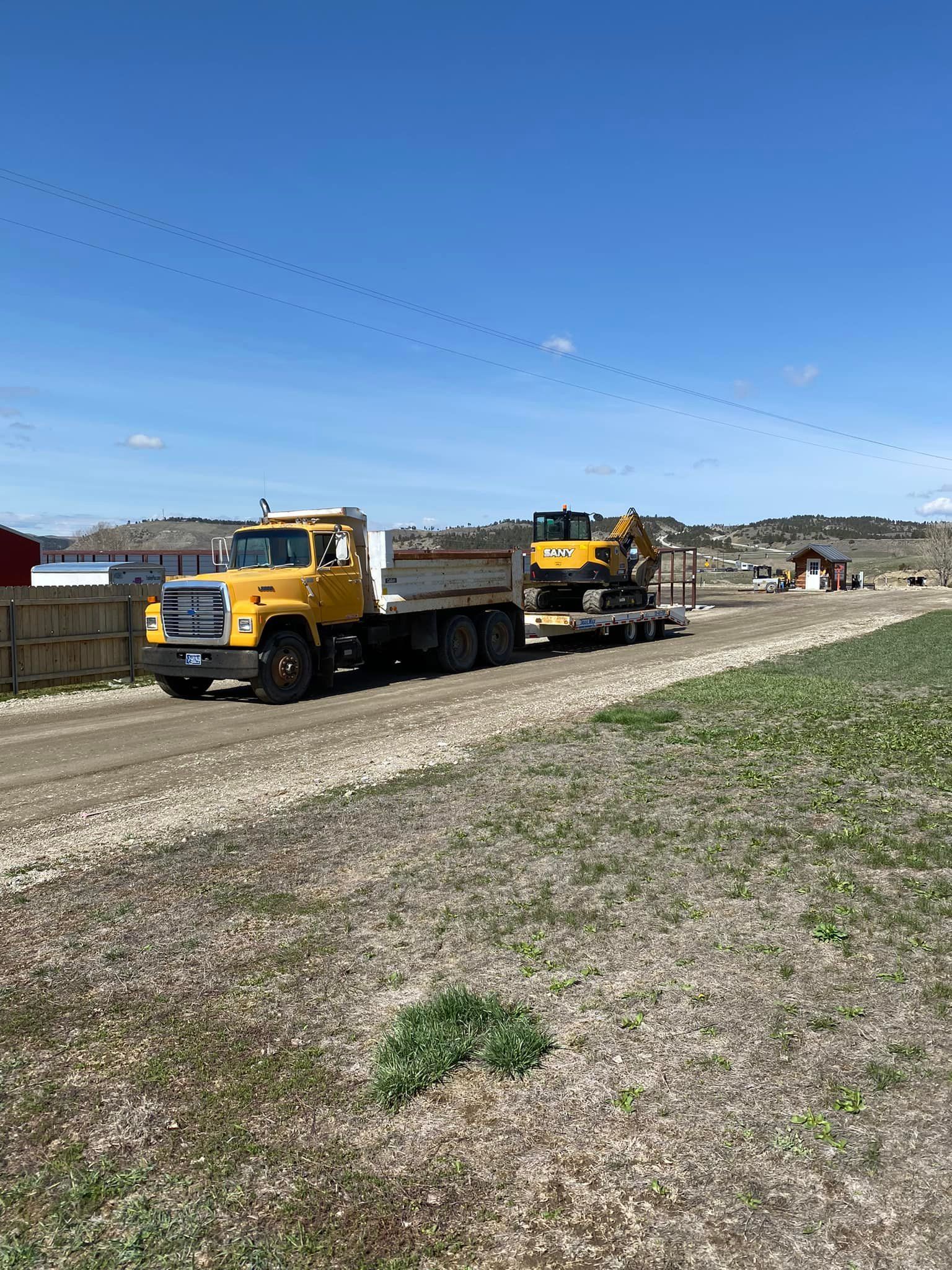 Yellow dump truck towing excavator on a gravel road, under a blue sky.