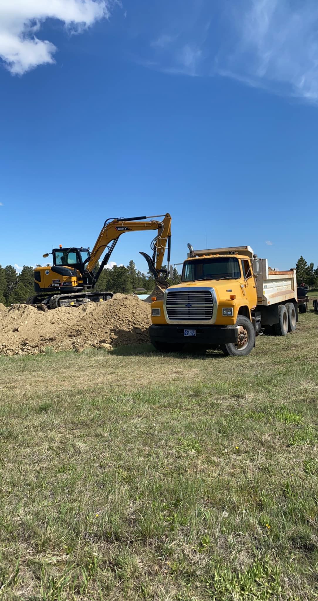 Yellow excavator loading dirt into a yellow dump truck in a field under a blue sky.