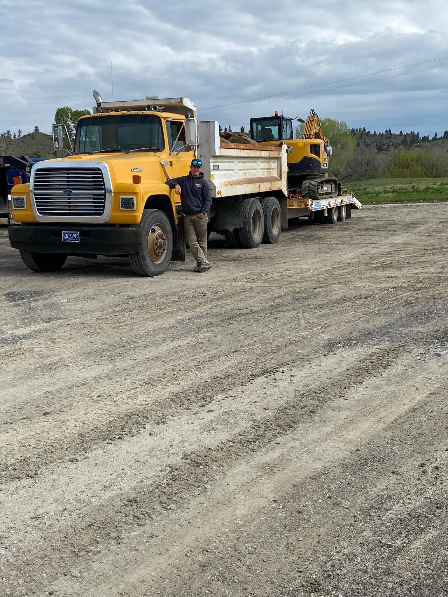 Yellow dump truck towing an excavator on a trailer; a man stands beside it on a gravel lot.
