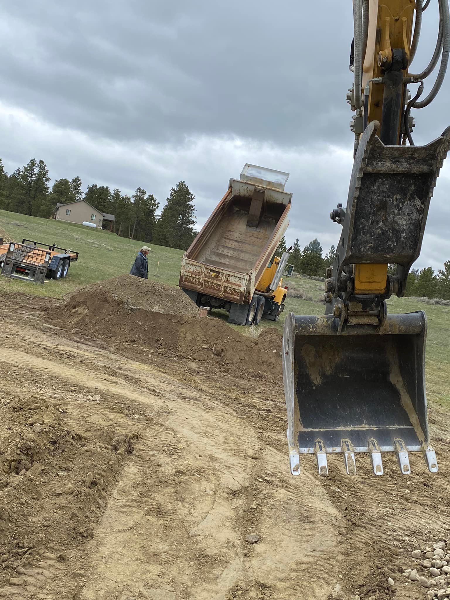 Excavator dumping soil into a truck on a hillside. Overcast sky, dirt and grass.
