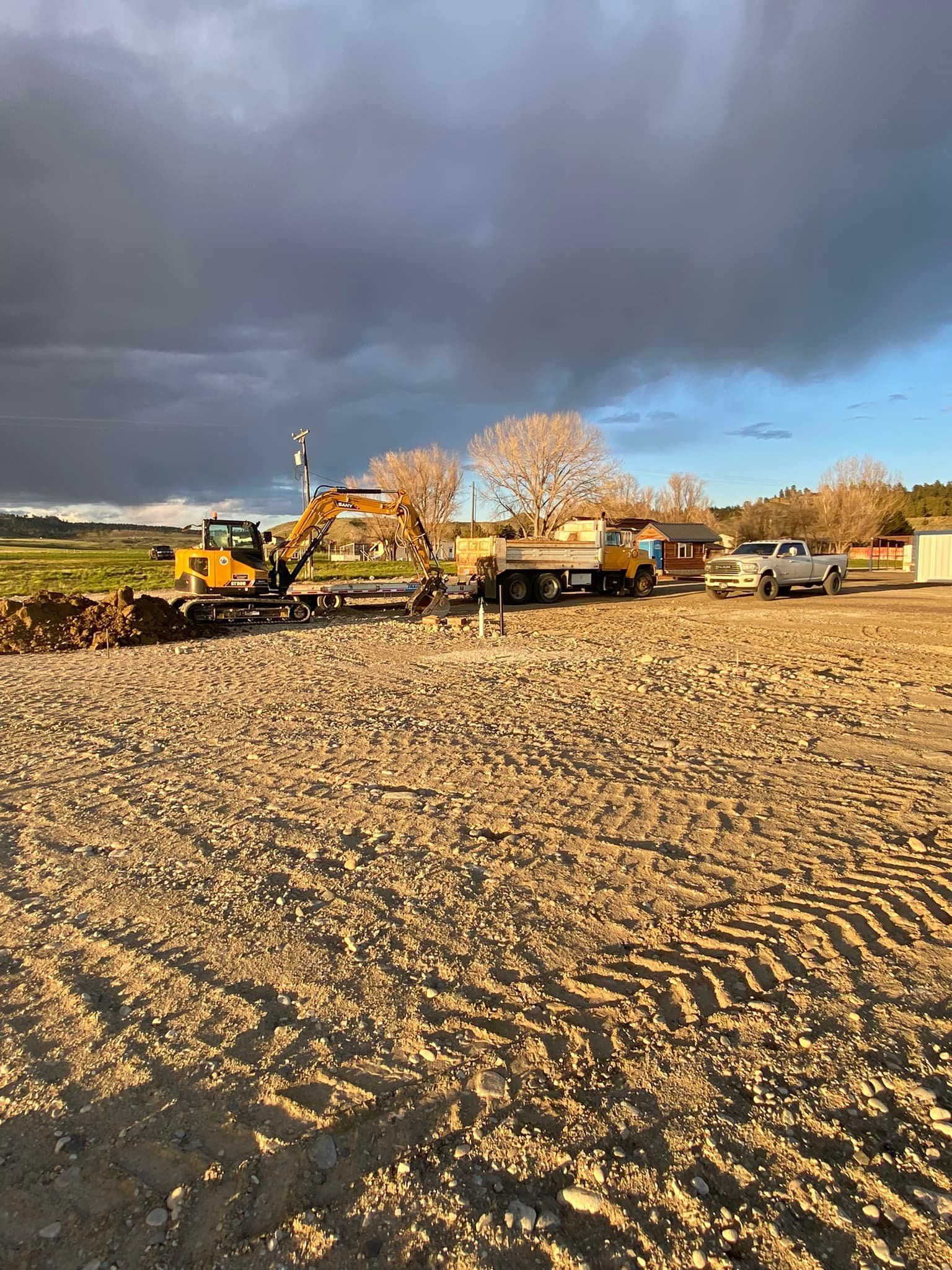 Construction site: excavator loading logs onto a flatbed truck on a dirt lot, cloudy sky.