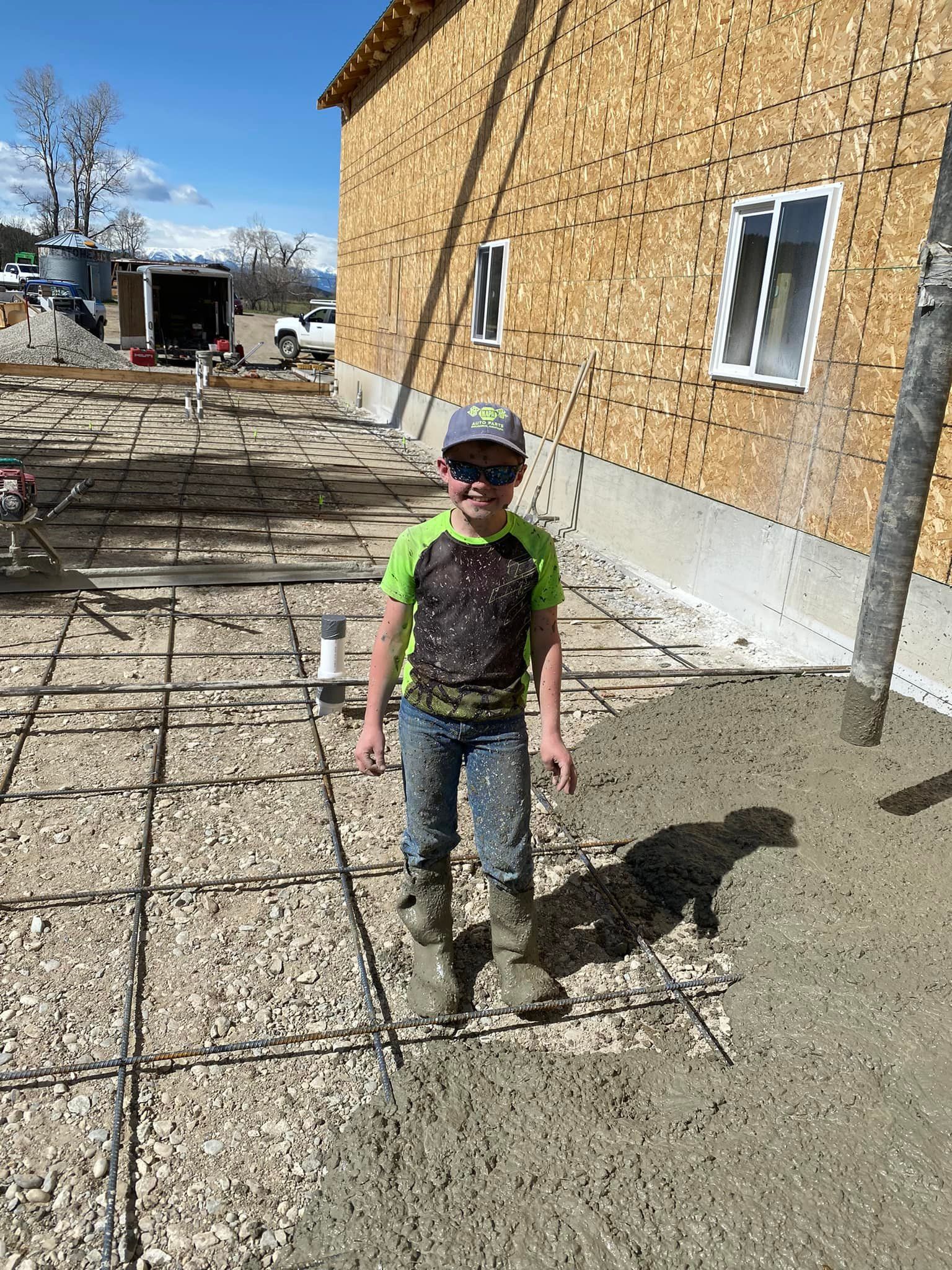Boy in hat and boots stands on rebar, next to concrete being poured for a building foundation.