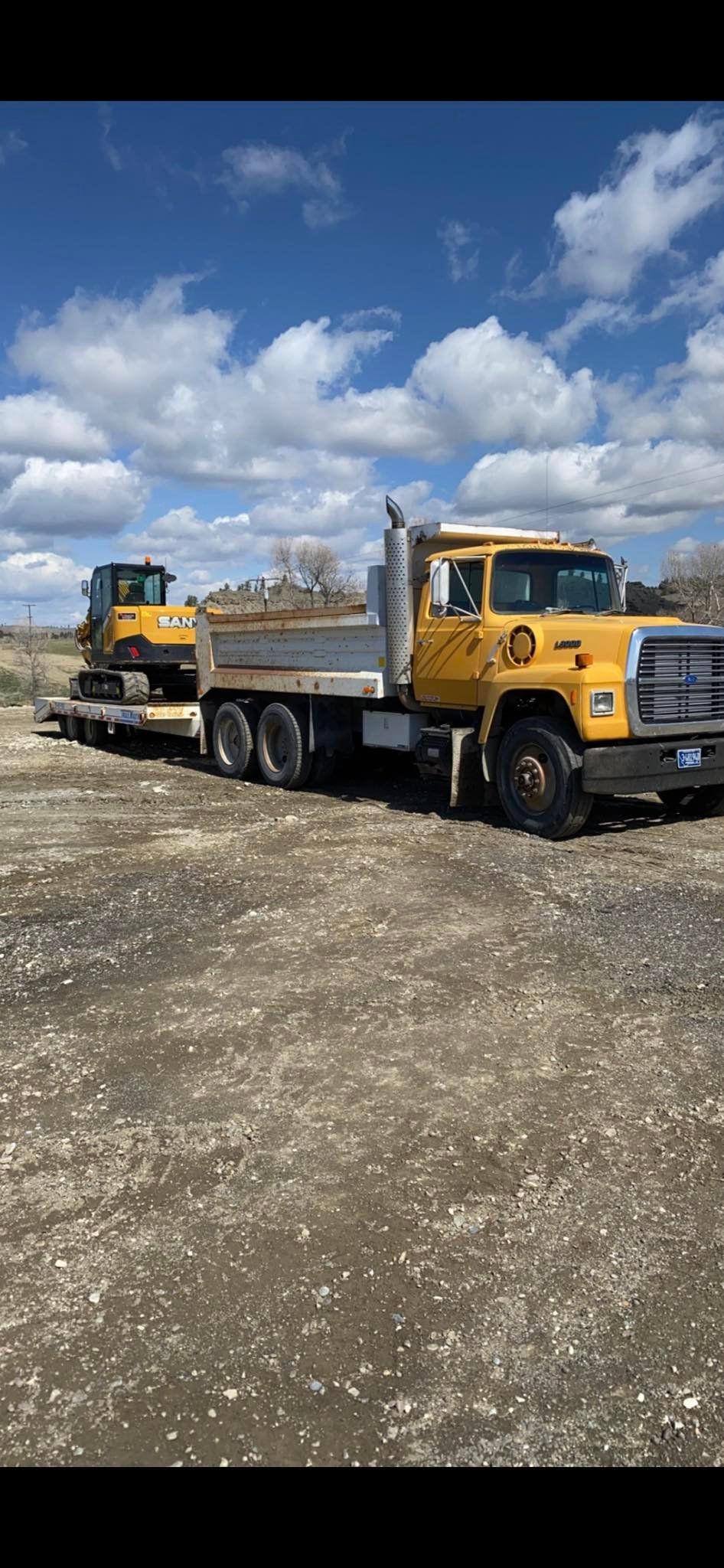 Yellow dump truck with a small excavator on a trailer, parked on a gravel lot under a blue sky with clouds.