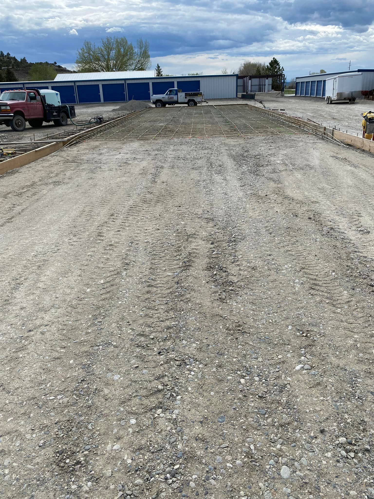 Gravel parking lot with bordering wood, small buildings, and a cloudy sky in the background.