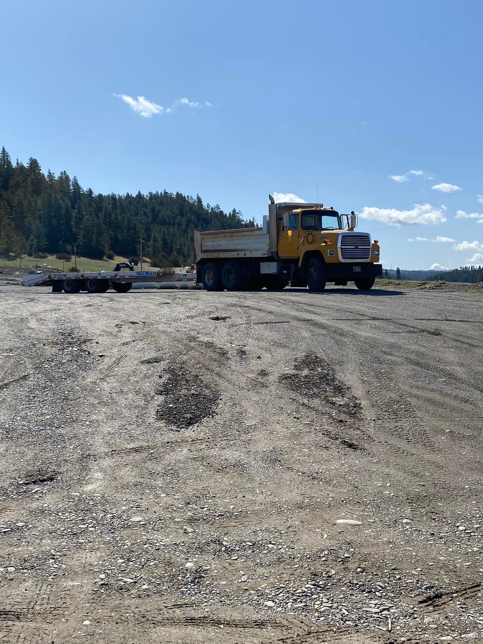 Yellow dump truck on a gravel surface. Trees and blue sky in the background.