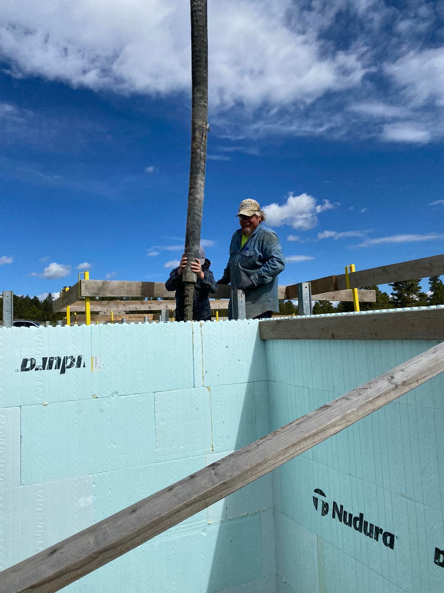 Two people working on a construction project, holding a concrete pouring tube against a blue insulated wall.
