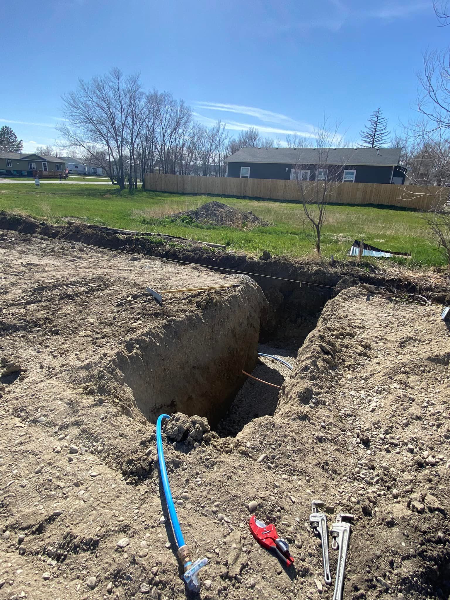 Trench dug in yard with blue water pipe, tools, and houses in the background under a blue sky.